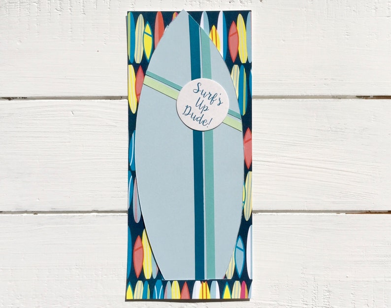 Op de afbeelding: Een blauw en wit surfplank met een gele streep en een ronde sticker met de tekst "Surf's Up Dude!" De achtergrond is een patroon van kleurrijke surfplanken.