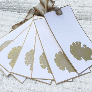 May include: White gift tags with gold turkey silhouettes and gold trim. A brown ribbon is attached to the tags. These tags are perfect for Thanksgiving or fall-themed gifts.