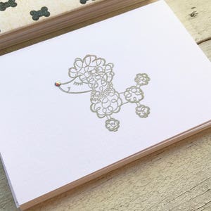 May include: A white card with a silver outline of a poodle with a rhinestone on its nose. The poodle is in a sitting position with its legs outstretched.