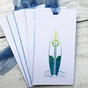 May include: A set of five white gift tags with blue trim, featuring a green and blue surfboard with a white flower design. The tags are tied together with a blue ribbon.