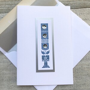 May include: A white card with a silver border and a blue and white illustration of a stylized figure with a hat and boots. The figure is made up of geometric shapes and has three silver circles on its body.