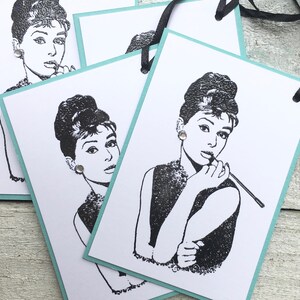May include: Three white gift tags with a black and white illustration of Audrey Hepburn. The tags have a light blue border and a black ribbon.