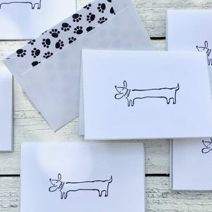 May include: Set of six blank greeting cards featuring a simple black and white illustration of a dachshund dog. The cards are white with black paw print envelopes.