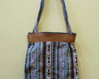May include: A brown leather tote bag with a blue, green and white patterned fabric body. The bag has two long straps.