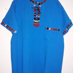 Puede incluir: Una camisa azul de manga corta con un cuello decorativo y un bolsillo en el lado izquierdo. El cuello tiene un ribete estampado en negro, rojo y blanco. La camisa tiene un detalle de encaje en la parte delantera.