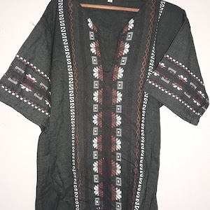 Puede incluir: Una túnica verde oscuro con cuello en V y bordados geométricos marrones y blancos. La túnica tiene mangas cortas y un corte holgado.