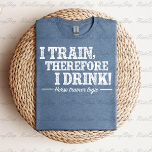 May include: A heather blue short-sleeve t-shirt featuring white distressed text that reads I TRAIN, THEREFORE I DRINK! and Horse trainer logic below it. The shirt has a crew neckline and is displayed folded on a round, woven, light brown textured surface.