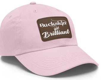 Könnte beinhalten: Eine rosafarbene Baseballkappe mit einem braunen Lederpatch, auf dem in weißen Buchstaben "Buckskin and Brilliant" steht.