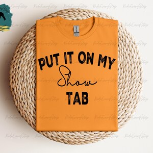K&ouml;nnte beinhalten: Orangefarbenes T-Shirt mit dem schwarzen Text "PUT IT ON MY Show TAB". Das Shirt liegt auf einem runden, gewebten Platzset. Ein Logo mit einer Pferdesilhouette ist oben links zu sehen.