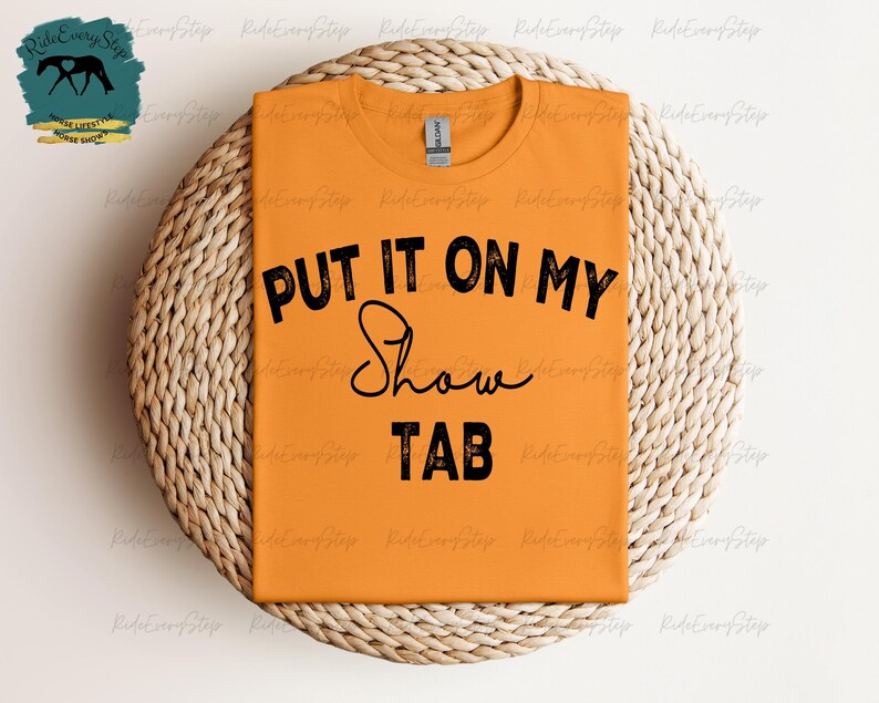 K&ouml;nnte beinhalten: Orangefarbenes T-Shirt mit dem schwarzen Text "PUT IT ON MY Show TAB". Das Shirt liegt auf einem runden, gewebten Platzset. Ein Logo mit einer Pferdesilhouette ist oben links zu sehen.