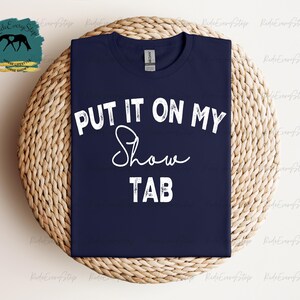 K&ouml;nnte beinhalten: Marineblaues T-Shirt mit dem wei&szlig;en Text "PUT IT ON MY Show TAB". Das Shirt liegt auf einer runden, gewebten Unterlage. Ein Logo mit einer Pferdesilhouette ist links oben sichtbar.