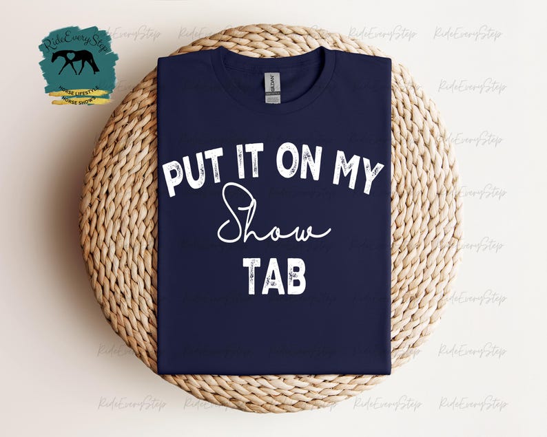 K&ouml;nnte beinhalten: Marineblaues T-Shirt mit dem wei&szlig;en Text "PUT IT ON MY Show TAB". Das Shirt liegt auf einer runden, gewebten Unterlage. Ein Logo mit einer Pferdesilhouette ist links oben sichtbar.