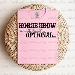 Könnte beinhalten: Ein hellrosa T-Shirt mit schwarzem Text im Used-Look, der HORSE SHOW WHERE SLEEP IS OPTIONAL lautet. Das Shirt hat einen Rundhalsausschnitt und einen klassischen Kurzarm-Schnitt und wird flach liegend mit sichtbarem Markenetikett am Kragen gezeigt.
