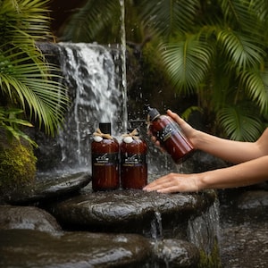 Könnte beinhalten: Drei braune Flaschen Kokosnuss-Shampoo und Duschgel werden in der Nähe eines Wasserfalls präsentiert. Die Flaschen haben schwarze Pumpen und sind mit Bindfäden und weißen Akzenten verziert. Die Etiketten tragen die Aufschrift "Coconut Lure".