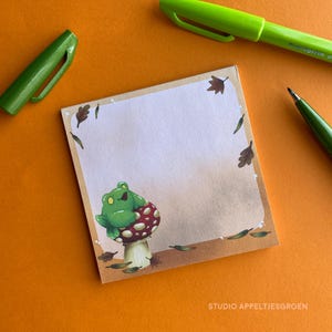 May include: A square notepad featuring a frog on a mushroom illustration. The notepad has a light brown border with leaves and a frog illustration. The text "STUDIO APPELTJESGROEN" is at the bottom. Two green pens are on the orange surface.