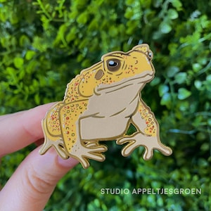 Könnte beinhalten: Eine goldfarbene Emaille-Anstecknadel in Form einer Kröte mit hellbeigem Bauch und einem strukturierten gelben Rücken mit orangefarbenen und braunen Flecken. Die Kröte hat ein markantes Auge mit dunkler Pupille und ausgeprägte Gliedmaßen mit gespreizten Zehen, eingefasst in eine goldene Metallkontur.