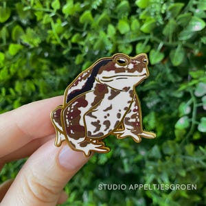 Könnte beinhalten: Ein Hartemaille-Pin in Form einer Kröte mit weißem Bauch und braunen, gefleckten Markierungen auf dem Rücken. Der Pin hat goldfarbene Metallumrandungen und eine glatte Oberfläche. Der Text STUDIO APPELTJESGROEN ist unten in Weiß gedruckt.