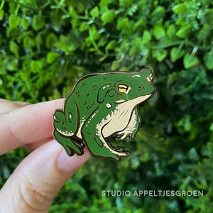 May include: A green and cream-coloured enamel pin shaped like a toad with a textured back, dark outlines, and yellow eyes. The pin features a detailed illustration of a sitting toad with a light-coloured underbelly and dark green skin markings.