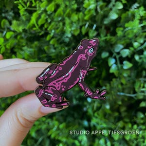 Könnte beinhalten: Eine dunkelviolette Emaille-Anstecknadel in Froschform mit leuchtend pinkfarbenen Mustern auf Körper und Gliedmaßen. Der Frosch ist im Seitenprofil mit einem sichtbaren Auge und detaillierten Umrissen dargestellt und hat eine glatte Oberfläche.