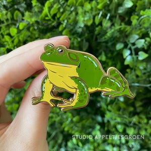 Könnte beinhalten: Eine Metall-Emaille-Anstecknadel in Form eines Frosches mit leuchtend grünem Rücken und gelber Unterseite. Der Frosch ist in sitzender Position dargestellt, mit detaillierten dunkelgrünen Flecken an Beinen und Rücken, umrandet von goldenem Metall.