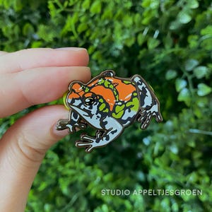 May include: A metal enamel pin shaped like a frog with a vibrant orange back, lime green patches, and black and white mottled markings. The frog is depicted in a crouching position with detailed outlines and a smooth, glossy finish on the surface.