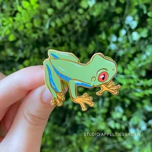 Könnte beinhalten: Ein grüner Frosch-Emaille-Pin mit blauen Streifen und goldenen Akzenten. Der Frosch hat rote Augen und gelbe Füße. Der Pin ist vor einem grünen Hintergrund platziert. Der Text "STUDIO APPELTJESGROEN" befindet sich unten.