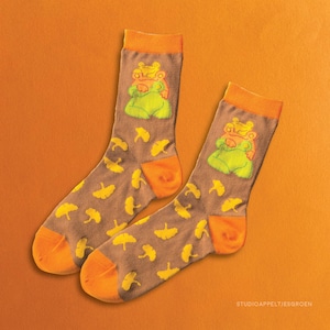 Può includere: Un paio di calzini marroni con un motivo a foglia di ginkgo giallo e una grafica di rana verde. I calzini hanno polsini arancioni.