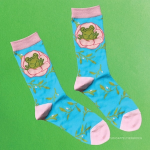 Puede incluir: Un par de calcetines azules con puños y punteras rosas. Cada calcetín presenta una rana verde dentro de una flor rosa. Los calcetines tienen un estampado de hojas verdes y puntos blancos. El texto "TUDIOAPPELTIESGROEN" es visible en el calcetín derecho.