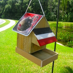 May include: A wooden bird feeder with a red, white, and black roof. The roof is decorated with a football helmet graphic. The feeder has a tray for birdseed.