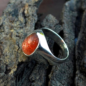 May include: A silver ring with a large, oval-shaped, red sunstone gemstone.