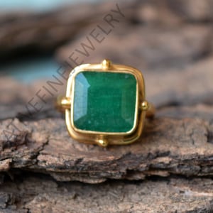 Puede incluir: Un anillo de oro con una piedra preciosa cuadrada de color verde esmeralda engastada en un engaste de bisel de oro. El anillo tiene un diseño sencillo y es perfecto para el uso diario.
