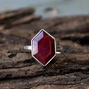 May include: A silver ring with a red gemstone set in a hexagonal shape. The ring is on a wooden surface.
