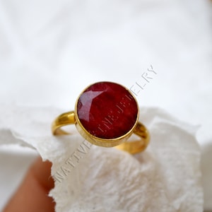 Könnte beinhalten: Ein goldener Ring mit einem großen, runden, facettierten Rubin-Edelstein. Der Rubin hat eine tiefrote Farbe und ist in eine goldene Lünette gefasst. Der Ring hat ein schlichtes, elegantes Design.