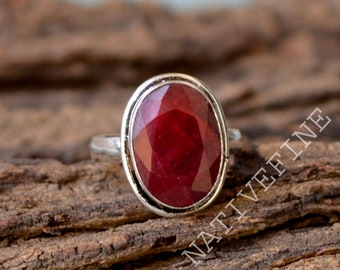 Op de afbeelding: Een zilveren ring met een grote, ovaal geslepen robijn edelsteen. De robijn heeft een dieprode kleur en is gezet in een zilveren bezel. De ring wordt gepresenteerd op een getextureerd, bruin houten oppervlak.