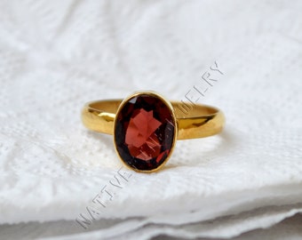 Puede incluir: Un anillo de oro con una piedra ovalada roja. El anillo está sobre una superficie blanca.