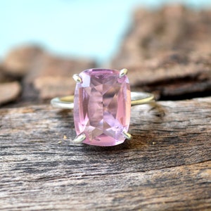 May include: A silver ring with a large, light pink, faceted gemstone. The ring is set on a piece of weathered wood.