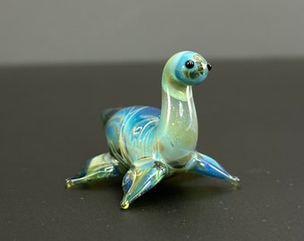 May include: A small glass figurine of a blue and green seal with black eyes. The seal is sitting on a black surface.