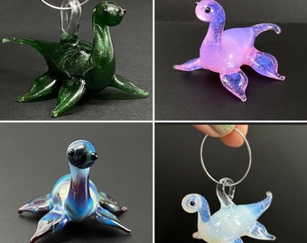 May include: Four glass ornaments shaped like a sea monster. The ornaments are green, pink, blue, and white. Each ornament has a loop at the top for hanging.