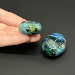 May include: Two glass art pieces resembling small, stylized bugs. The pieces are oval-shaped with a mix of blue, green, and white colors. Each has small, black, round eyes. One is held in a hand, the other rests on a dark surface.