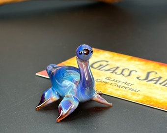 May include: A small, hand-blown glass sculpture of a blue and purple sea serpent with red accents. The serpent has a long neck and a small head with black eyes. The sculpture is sitting on a business card that reads "Glass Sale, Glass Art, Kate Corticelli."