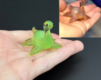 May include: A green glass sea serpent figurine with black eyes. The figurine is sitting in a hand.