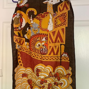 Puede incluir: Textil vintage marrón con una colorida ilustración del Arca de Noé. El diseño incluye animales, un barco y olas estilizadas en tonos naranja, amarillo y rojo. El textil está colgado de una percha.