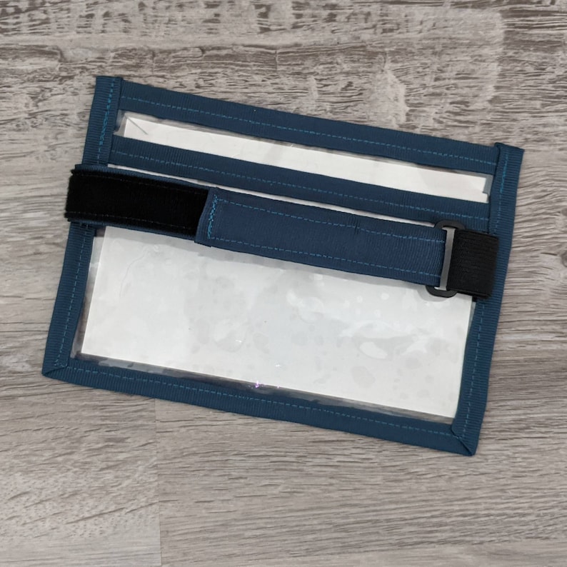 May include: A blue fabric pouch with a clear plastic window and a black adjustable strap. The pouch is designed to hold a small item, such as a phone or ID card.