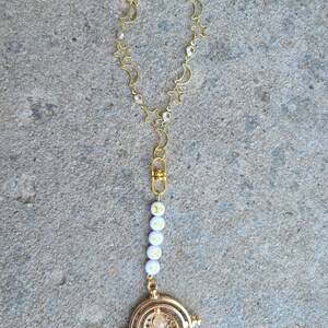 May include: A gold-coloured necklace with a charm of a time-turner from the Harry Potter series. The necklace has a chain with stars and crescent moons. The charm is attached to a string of white beads with the letters "ALANA" spelled out.
