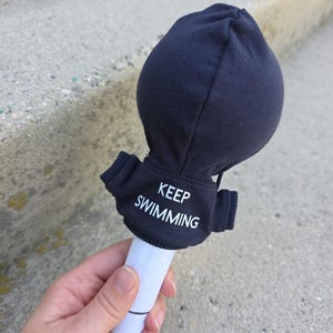 May include: A black fabric cover for a light stick featuring the white text KEEP SWIMMING printed on the front. The cover has a rounded top and small side sleeves, and it is fitted over a white cylindrical handle with a thin black line near the base.