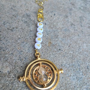 May include: A gold necklace with a white beaded charm that spells "ATINY" and a gold Time-Turner pendant with a clear hourglass inside.