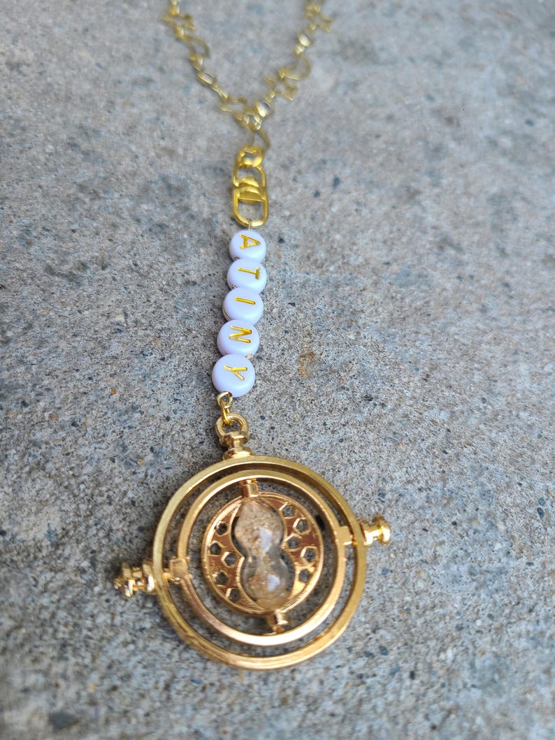 May include: A gold necklace with a white beaded charm that spells "ATINY" and a gold Time-Turner pendant with a clear hourglass inside.