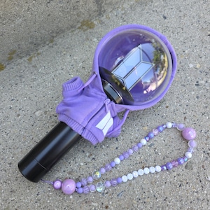 Pode incluir: Um bastão de luz preto com um topo esférico transparente contendo um logotipo BTS, vestido com um moletom roxo com capuz e um pequeno logotipo branco. Uma pulseira de contas em tons de roxo e branco com o texto BANGTAN está no cabo.