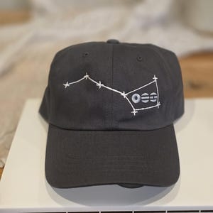 Puede incluir: Gorra de béisbol gris carbón con un diseño de constelación bordada en blanco y las letras "OSS". La gorra tiene una visera curva y una construcción clásica de seis paneles. La gorra está sobre una superficie blanca.