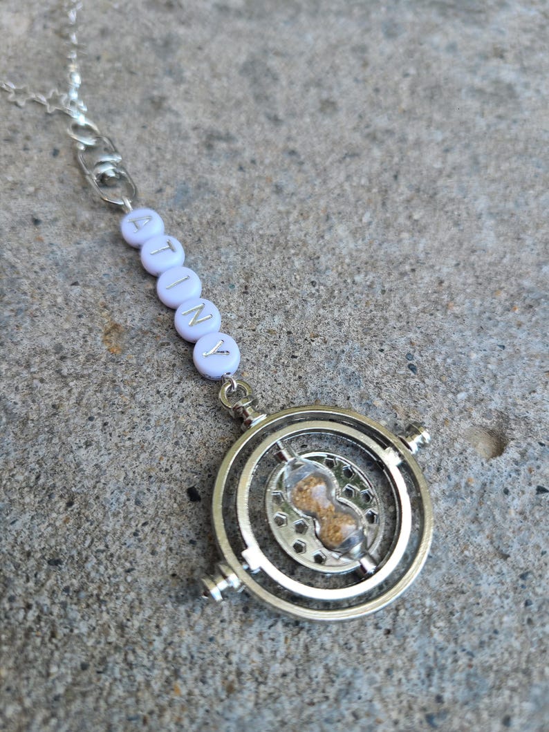 May include: A silver necklace with a white beaded charm that spells "ATINY". The charm is attached to a silver hourglass pendant.